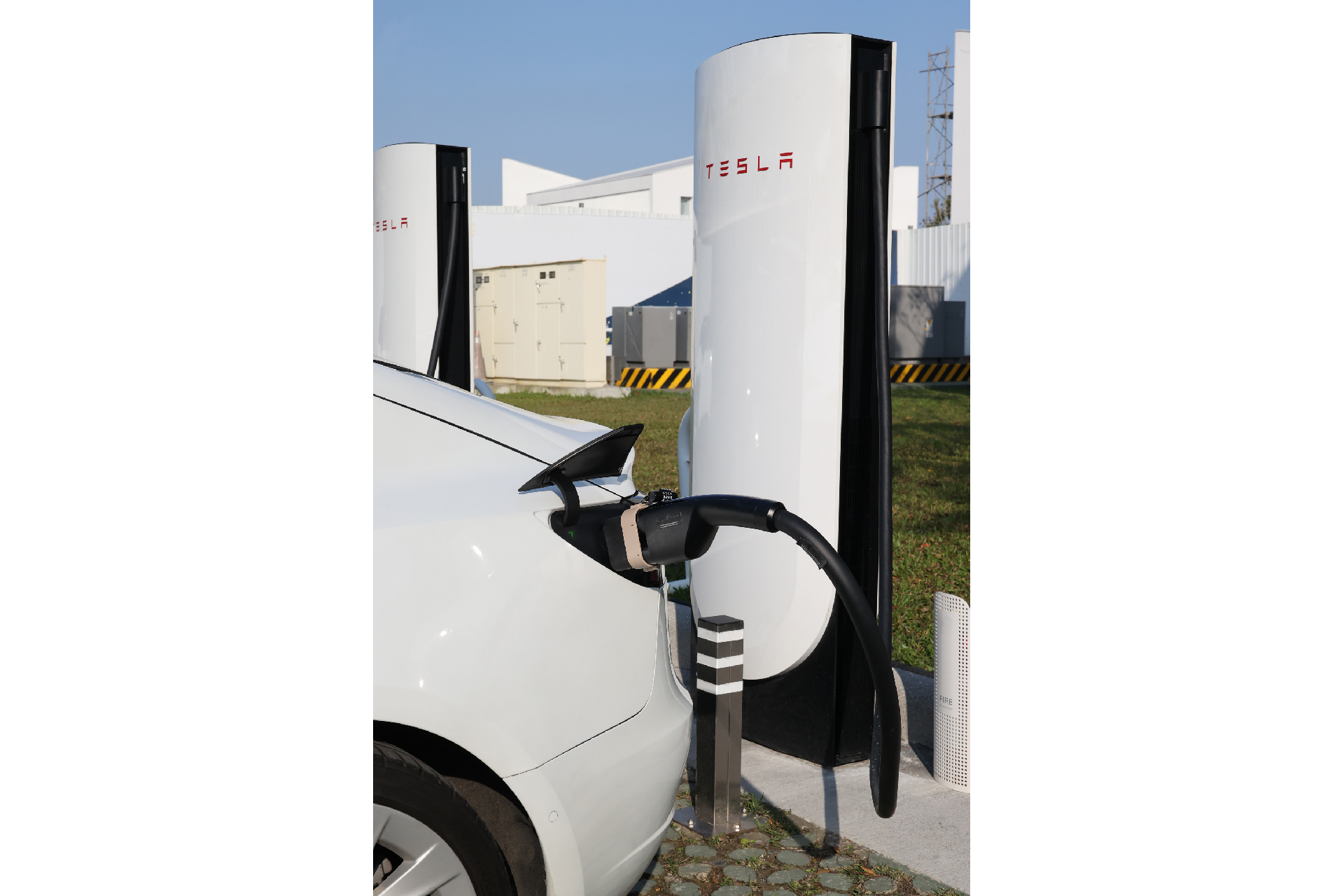 一輛白色 Tesla 在戶外的 Supercharger 充電，連接 JOWUA Taiwan CCS2 to NACS (TPC) 直流轉接頭；場景包括鋪好的地面、草地和背景中的建築物。.