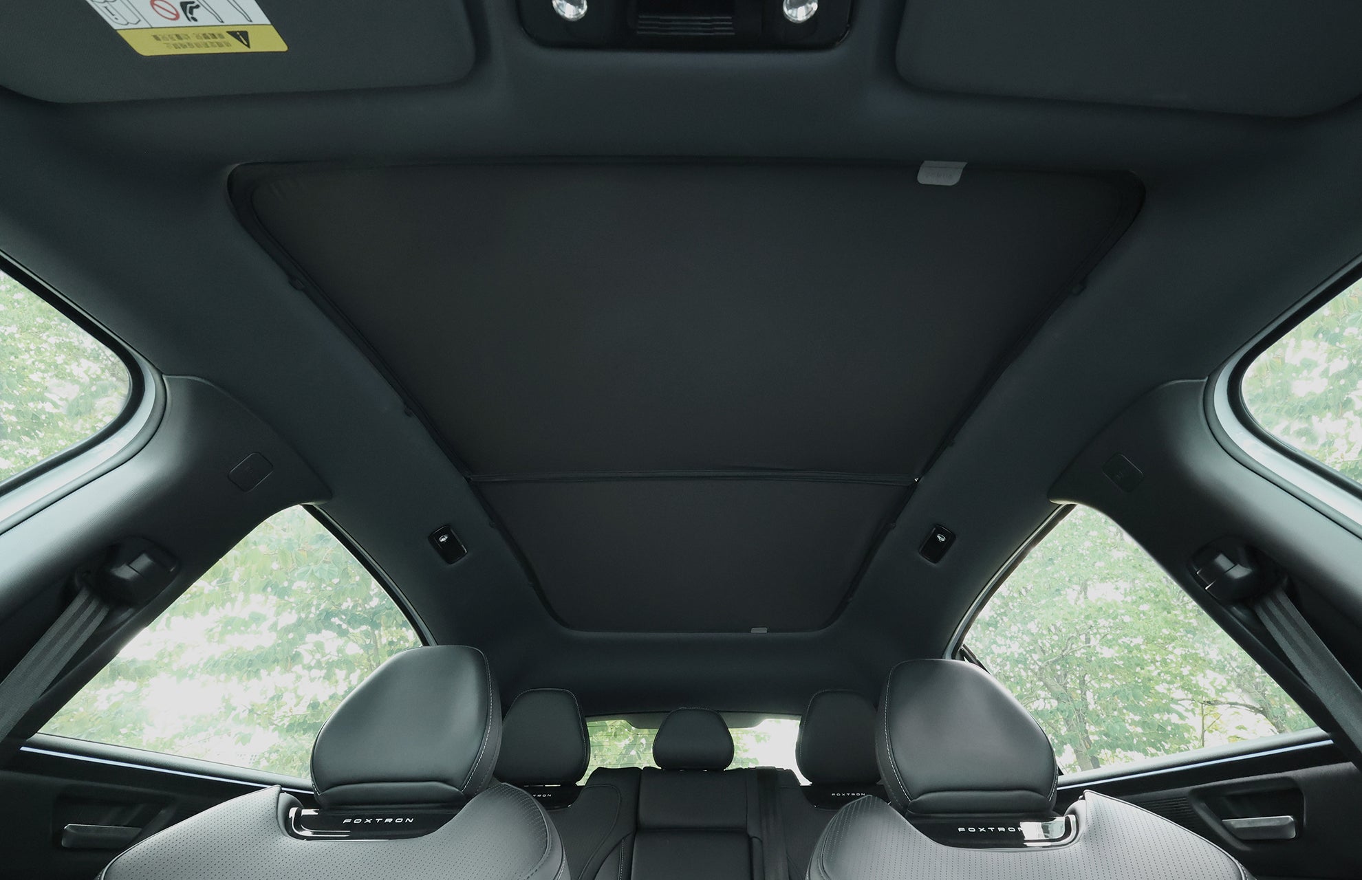 從後排座位，看到由台灣 JOWUA 玻璃車頂遮陽簾 (Foxtron Bria) 所覆蓋的封閉式全景天窗，可防 99% 的紫外線。車內有灰色真皮座椅、大車窗，車外有綠色風景。.