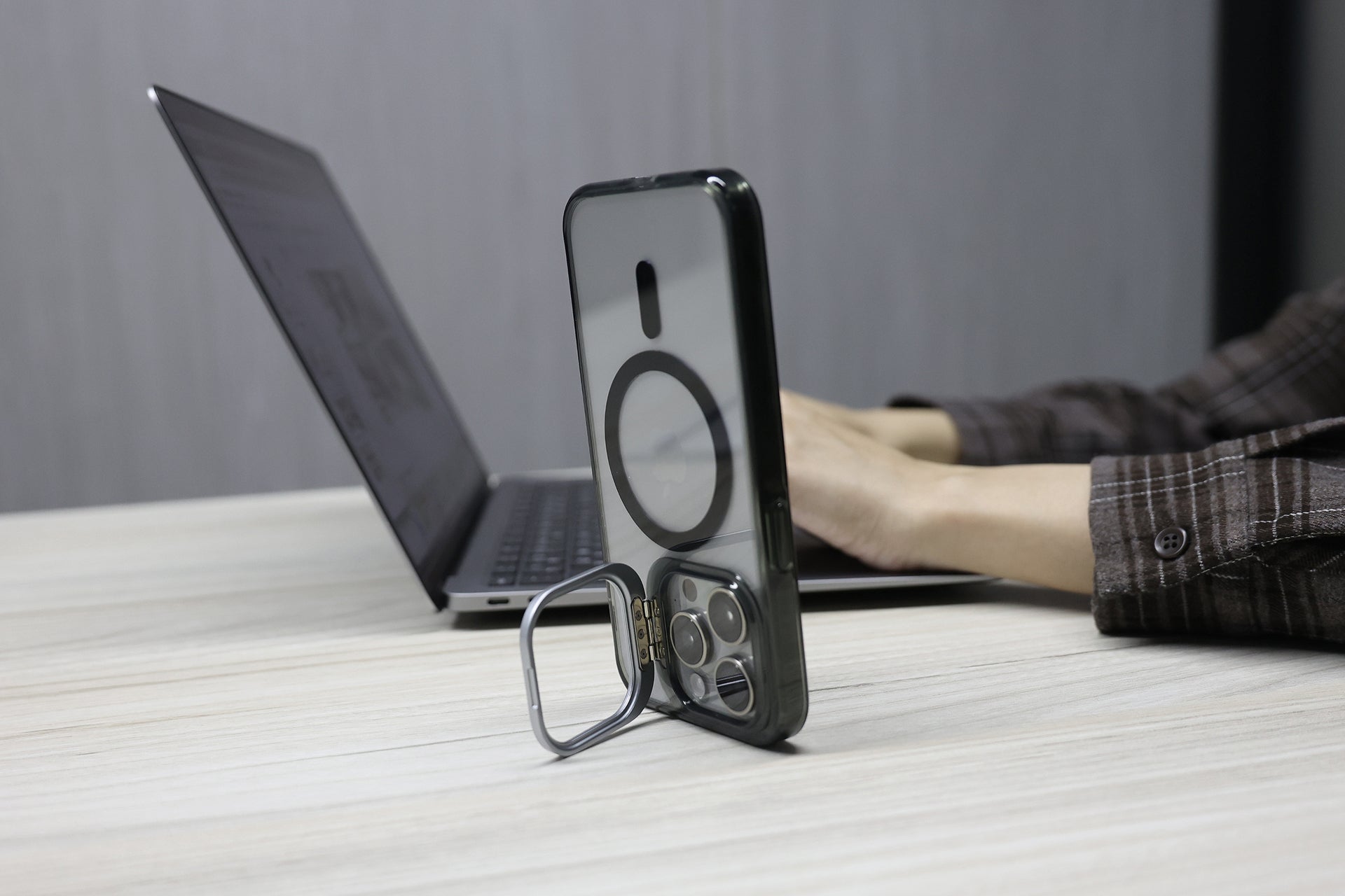 一個人在輕巧的辦公桌前使用筆記型電腦打字，而一隻裝有 JOWUA Taiwan MagSafe 支架 透明保護殼 (iPhone 15/14/13 系列) 的 iPhone 就支撐在附近。.
