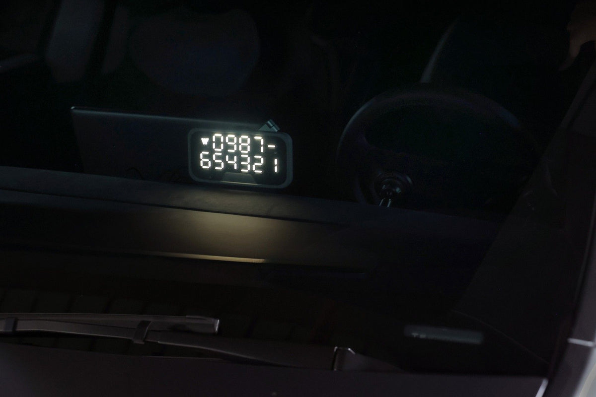 一款 JOWUA 台灣萬用 MagSafe 磁吸燈，外型酷似「臨時停車」電話卡，上面顯示著數字 0987 和 654321，放置在汽車儀表板上——透過擋風玻璃可見，背景中可見方向盤。.