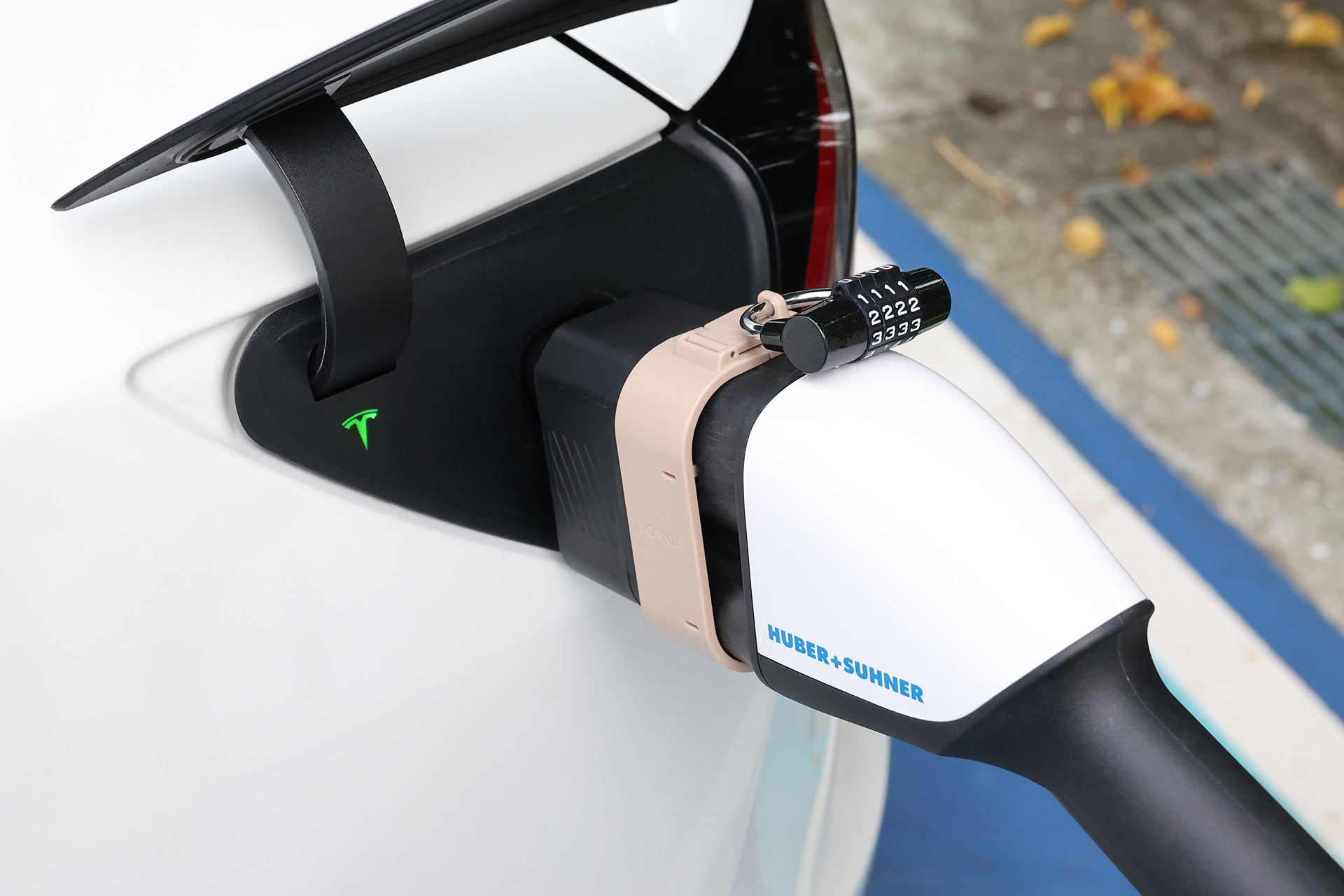 一輛白色 Tesla 在充電站使用 JOWUA Taiwan CCS2 至 CCS1 充電防拔鎖扣，充電站內有 HUBER+SUHNER 充電器，並以密碼鎖固定。綠色指示燈清晰可見，背景是樹葉和排水柵。.