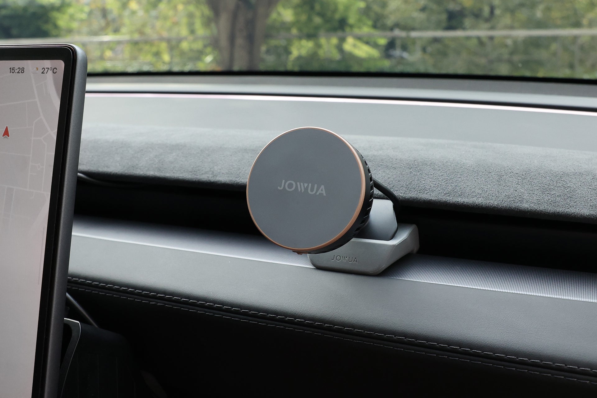 一台 JOWUA Taiwan Qi2 製冷出風口 MagSafe 車架 (Model 3/Y 煥然一新版) 安裝在觸控螢幕顯示器旁邊，透過擋風玻璃可以看到樹木和綠地。.