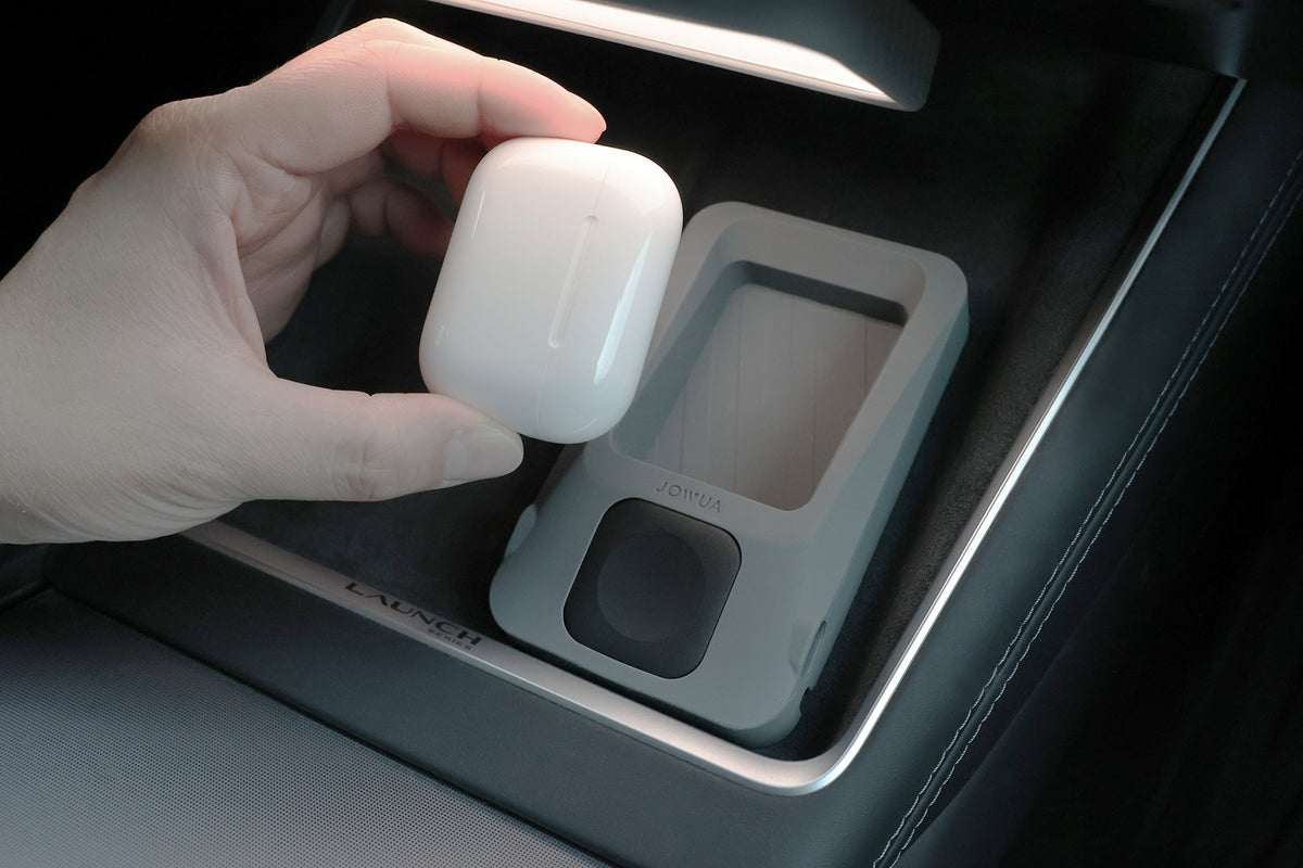 一隻手握著白色無線耳機充電盒（例如 AirPods 無線充電盒），置於汽車中控台上方，該中控台上擺放著 JOWUA 台灣多功能無線充電底座＋智慧手錶無線充電器，以及一個標有 JOYEA 字樣的托盤。.