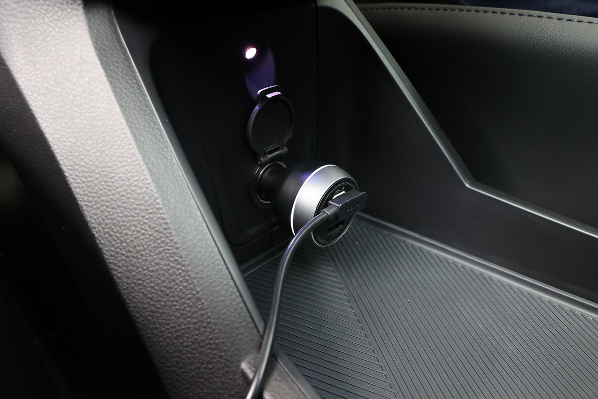 一幅汽車內飾的特寫畫面顯示，一個 USB 充電器插在 12V 插座上，充電線一端連接著 JOWUA 台灣製的 Qi2 製冷六維隱藏車架 (MagSafe 款)｜Foxtron Cavira 專用，該車架同時具備 Qi2 無線充電與 MagSafe 磁吸充電功能。.