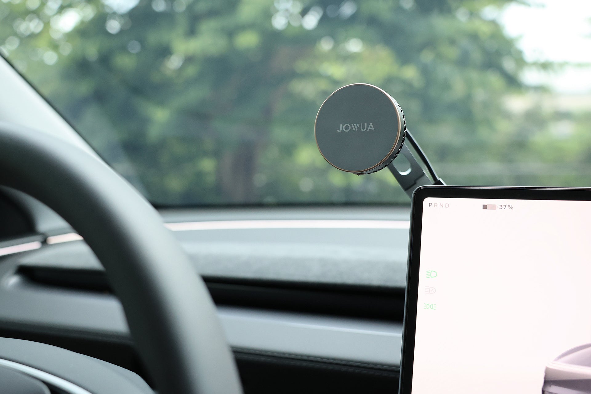 JOWUA Taiwan Qi2 製冷六維隱藏車架 (MagSafe 款) 安裝在儀表板上的數位顯示器旁邊。汽車的方向盤和擋風玻璃清晰可見，背景是模糊的樹木。.