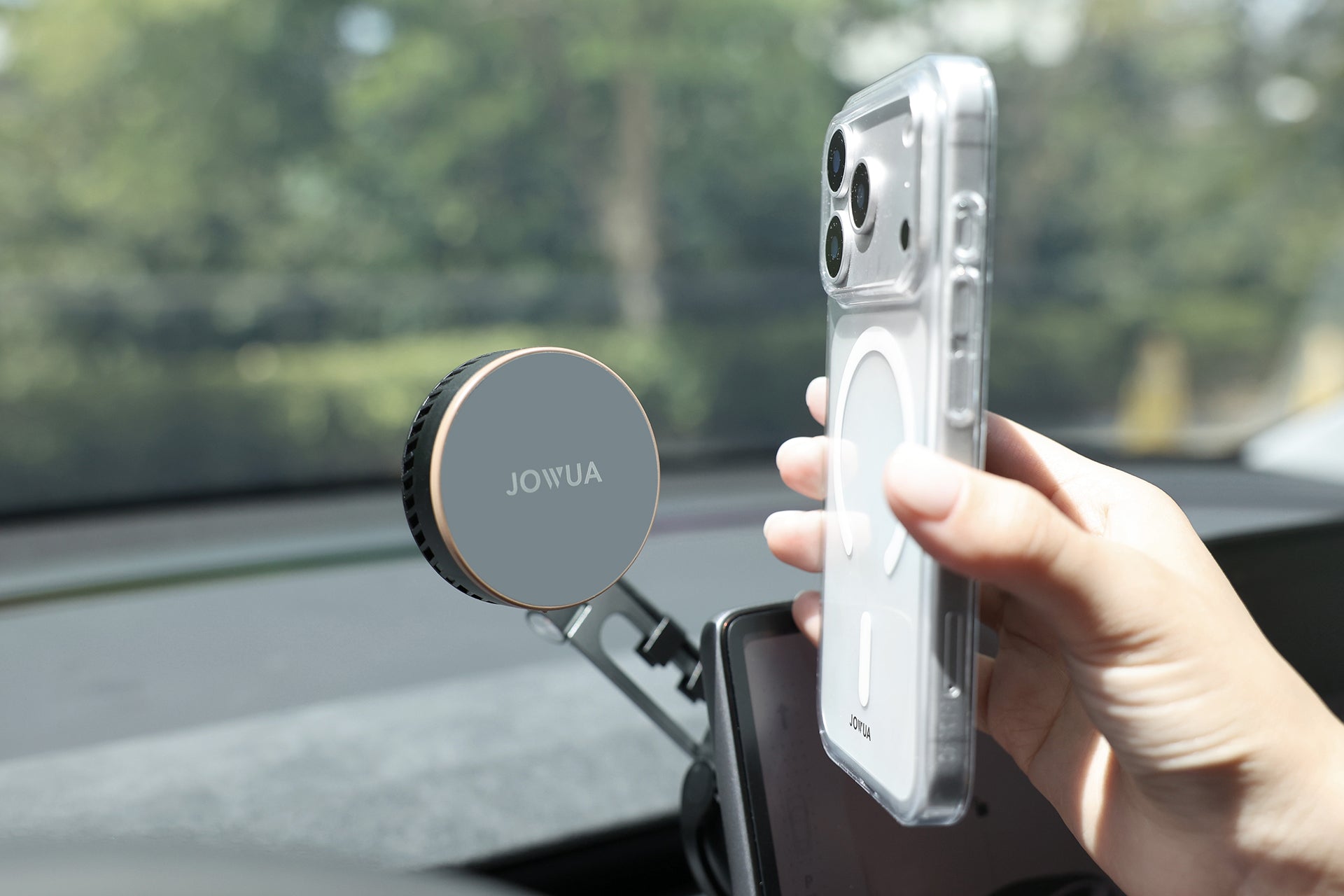 一隻手拿著智慧型手機，靠近汽車儀表板上的 JOWUA Taiwan Qi2 製冷六維隱藏車架 (MagSafe款)。窗外明亮的綠意突顯出這個冷酷的隱藏式 Tesla 手機支架，提供無縫的便利性。.