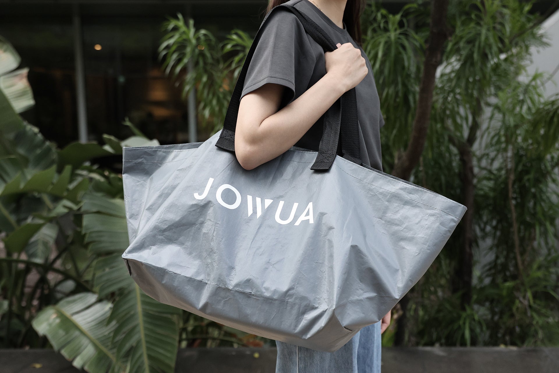 一個穿著深色 T 恤和牛仔褲的人，拿著一個大的灰色 JOWUA Taiwan 35L 環保購物袋，站在戶外的綠色植物旁。.