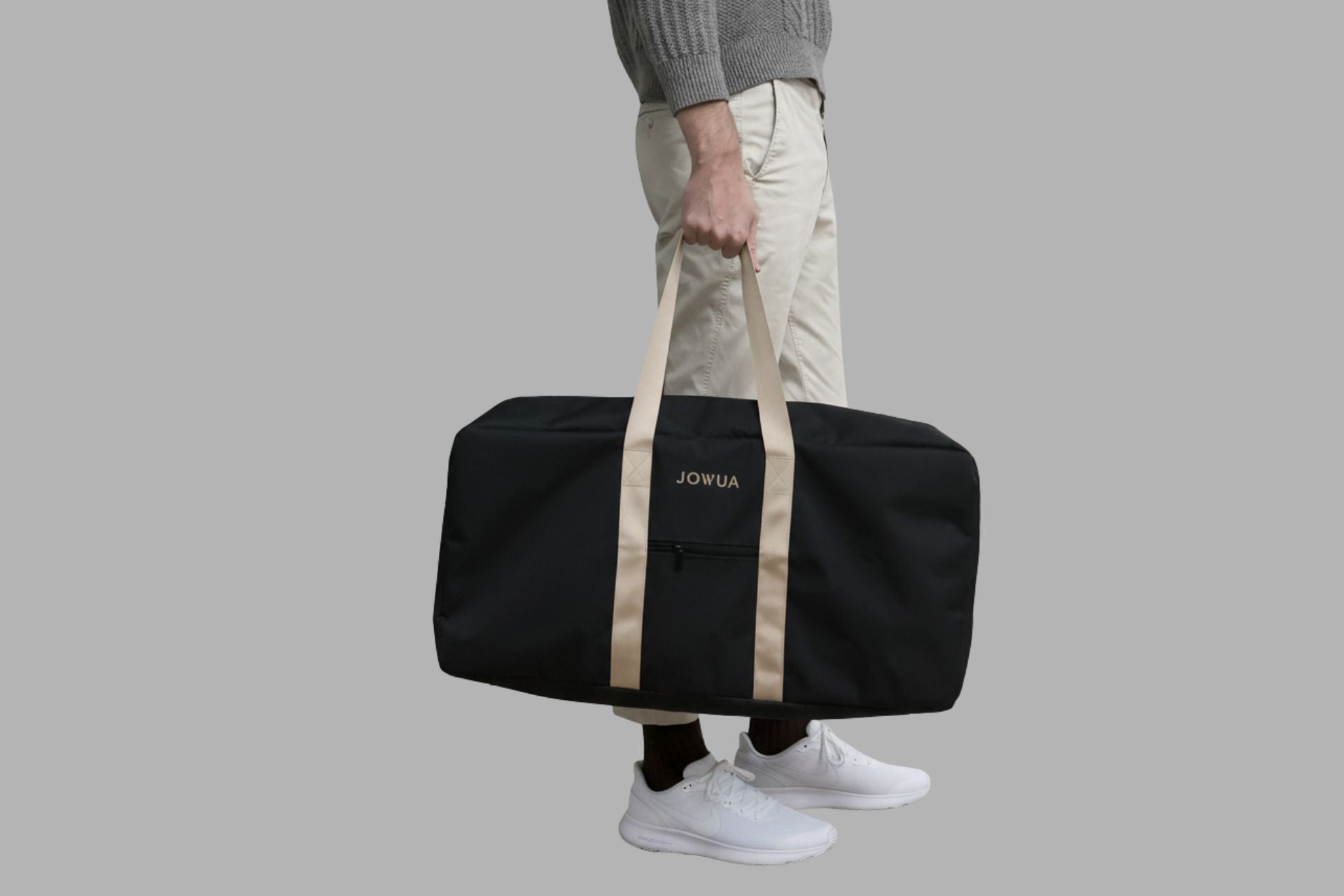 一個身穿灰色毛衣、米色褲子和白色運動鞋的人拿著 JOWUA Taiwan 前車廂 & 滑板車專用收納袋 (33L大容量)，搭配米色提手，是理想的特斯拉前車廂收納。流線型的包包在純灰色背景中顯得格外突出。.