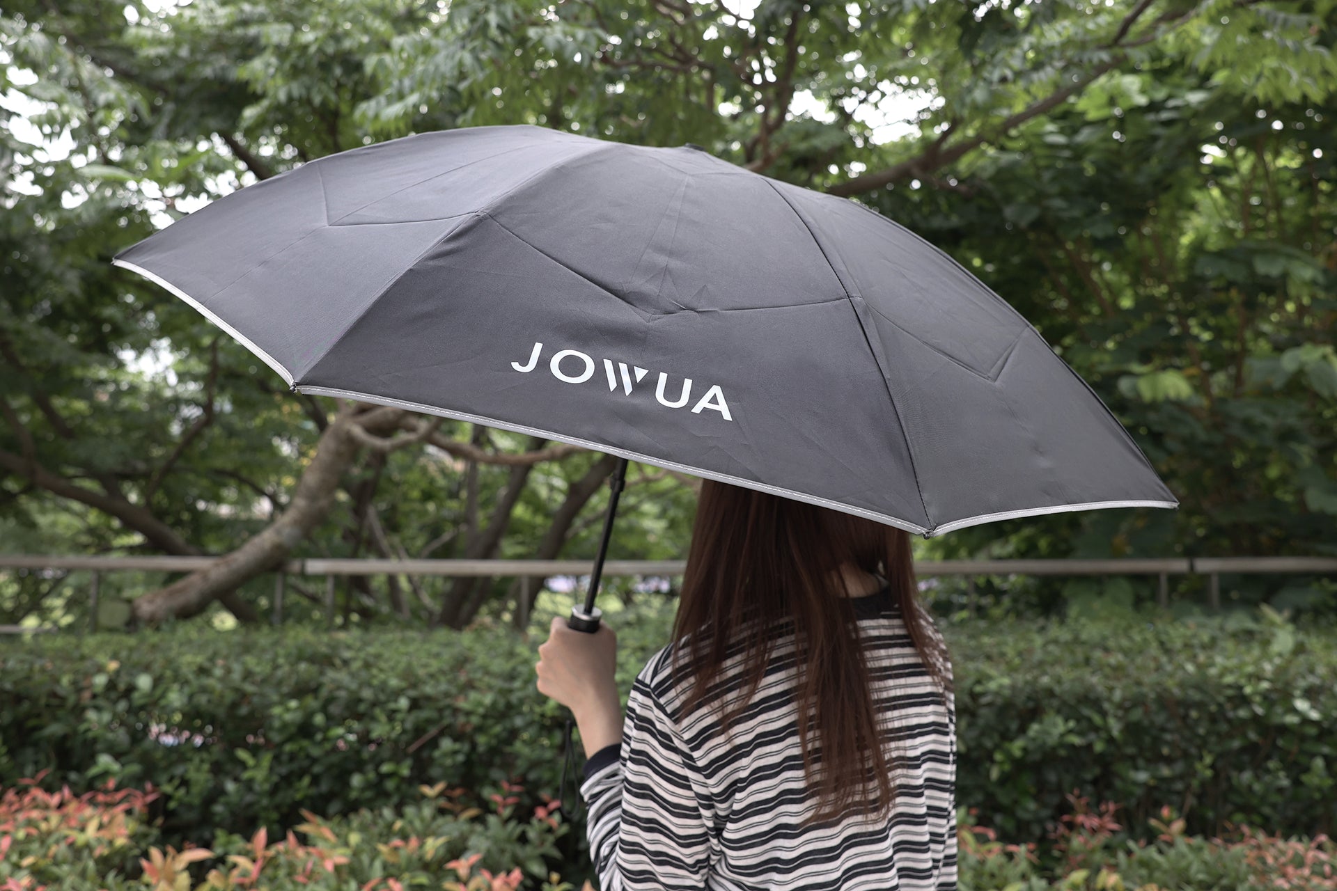 一位棕色長髮、身穿條紋襯衫的人拿著 JOWUA Taiwan 一鍵開收電動傘，站在外面的綠色灌木叢和樹木附近。.