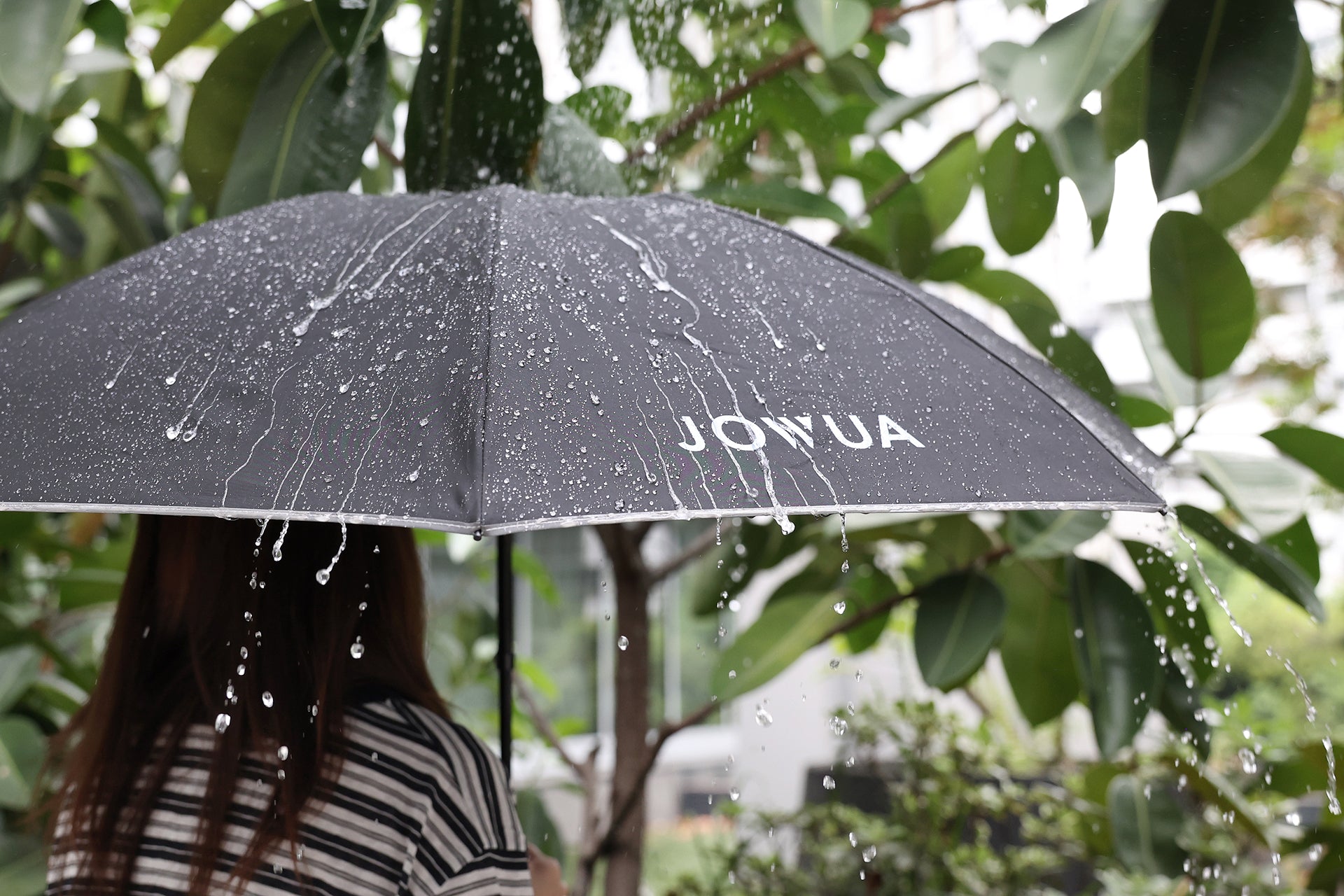 一個人拿著黑色的 JOWUA Taiwan 一鍵開收電動傘，站在雨滴與綠葉下，模糊的戶外背景。.
