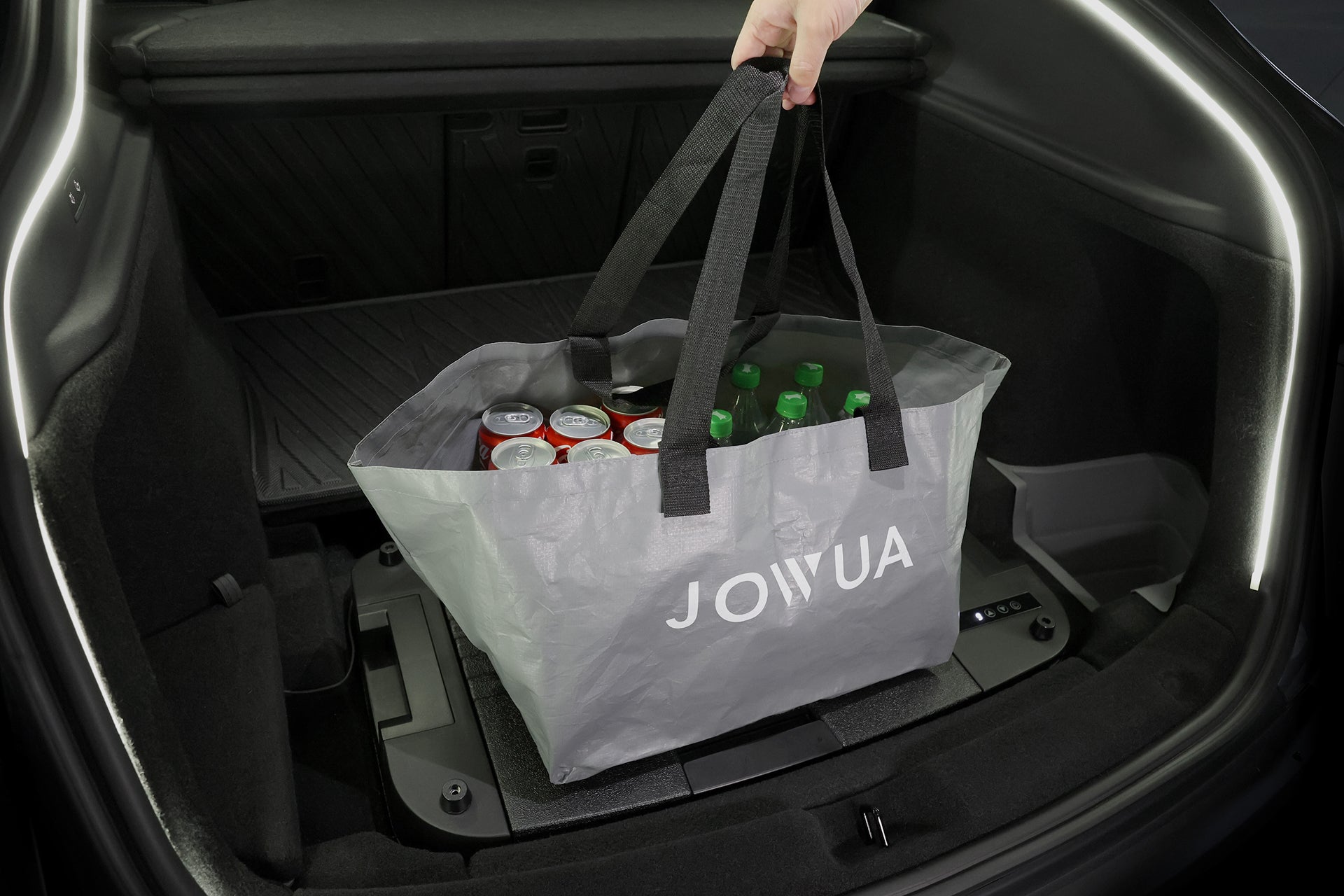 一隻手從車箱中提起裝滿飲料的灰色 JOWUA 可重複使用袋子，搭配您的 JOWUA Taiwan Type-C 車用行動冰箱 (Model Y全車系)，讓您的旅途更便利。.