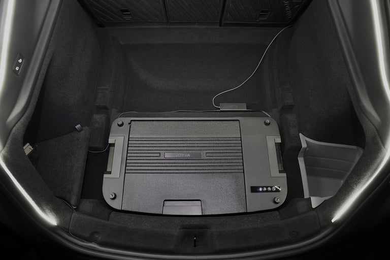 將容量為 35L 的 JOWUA Taiwan Type-C & 點菸器 雙軌車用行動冰箱 (Model Y全車系) 置於鋪有深色地毯的汽車後備箱中央，並將充電線插入右側的裝置。.
