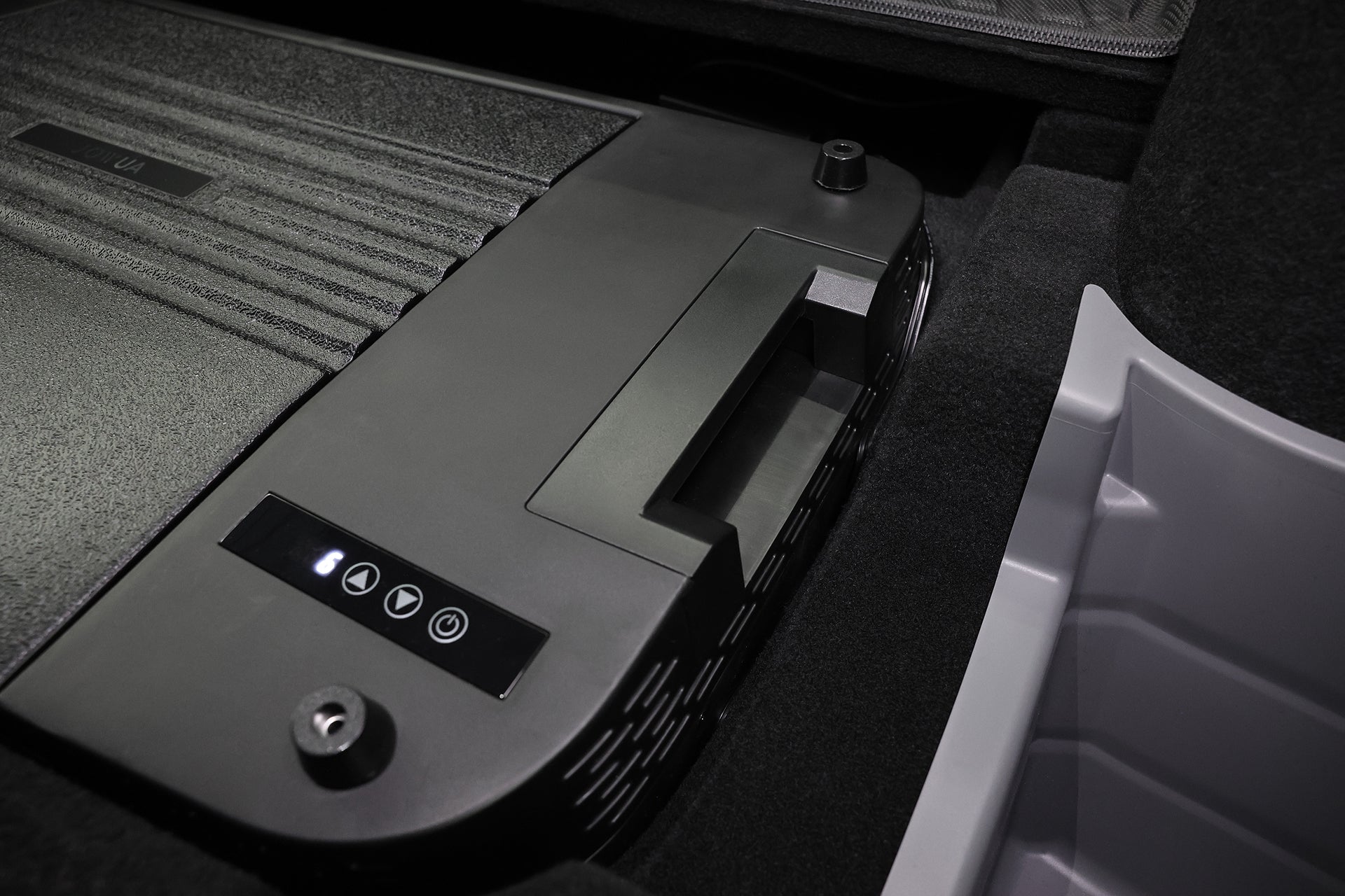 黑色的 JOWUA Taiwan Type-C 車用行動冰箱 (Model Y 全車系) 特寫，有控制按鈕和紋理表面，放置在淺灰色塑膠垃圾桶旁，擺放在深色地毯區域。.