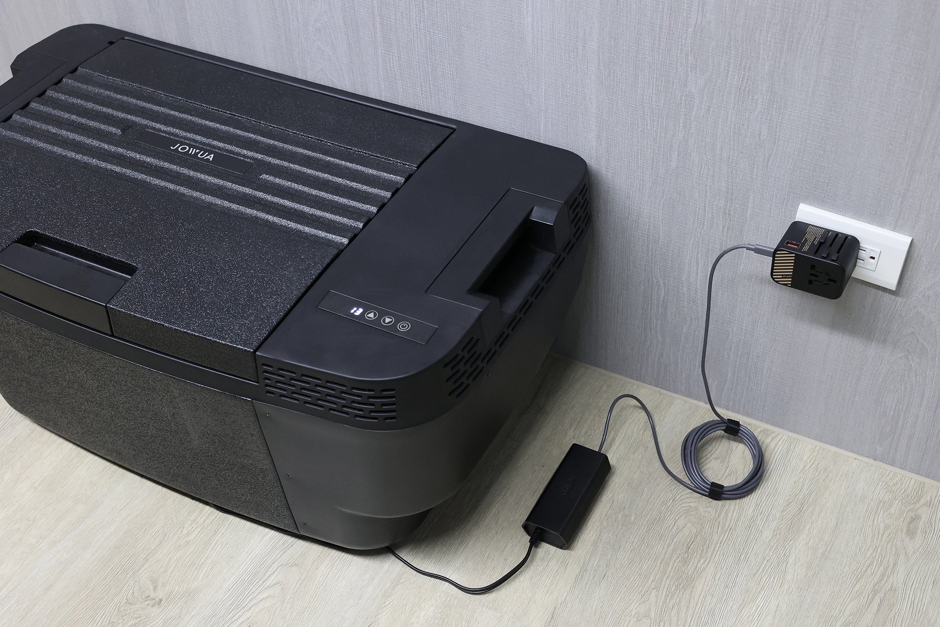 一台黑色的 JOWUA Taiwan Type-C 車用行動冰箱 (Model Y 全車系) 透過轉接器和電源線插入牆上的插座，放置在灰色牆壁旁的淺色木地板上。.