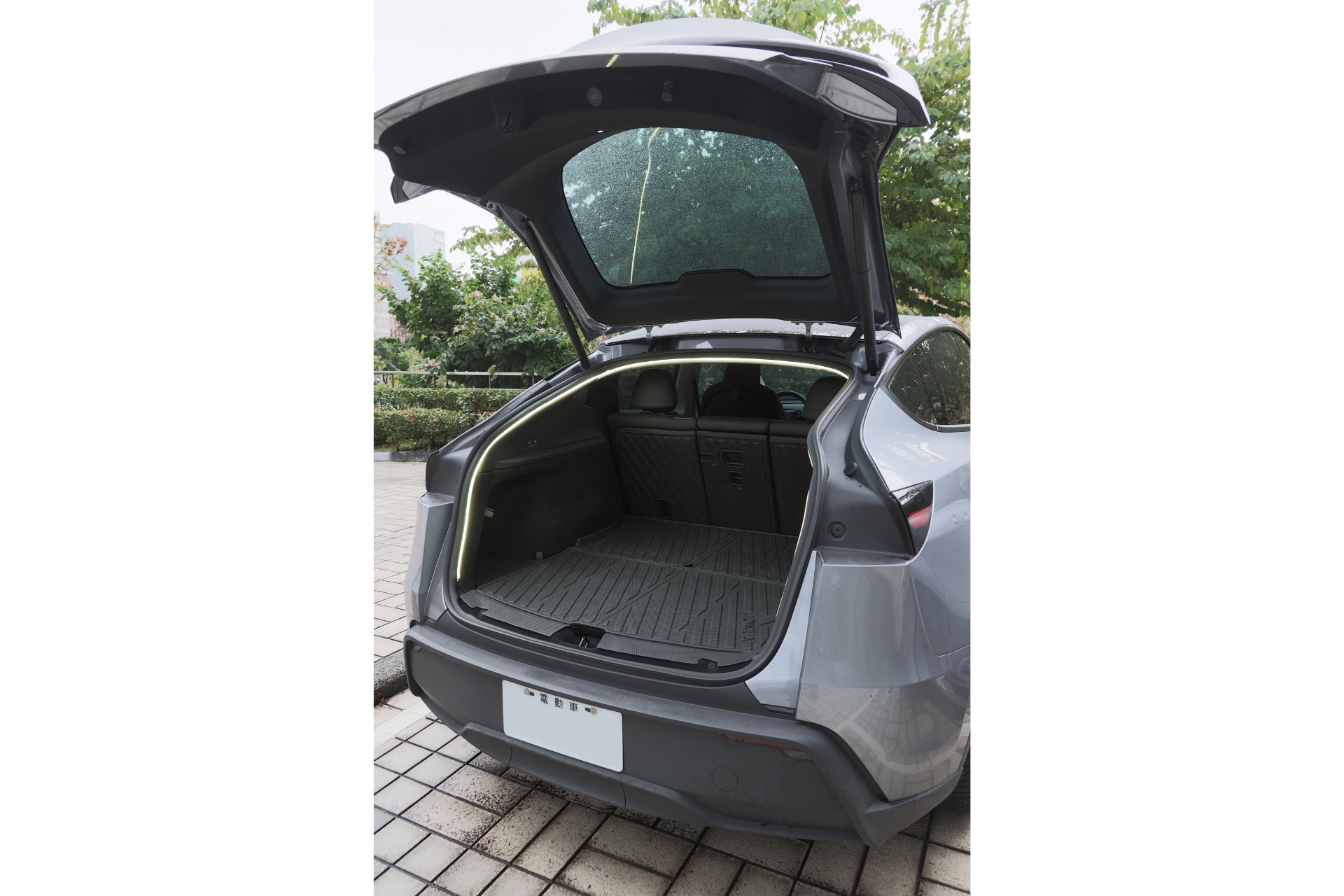 銀色的休旅車，打開後車艙門，展現寬敞的後車廂。搭配 JOWUA Taiwan 後車廂氣氛燈｜真言套索，提升 Model Y 的格調。樹木與地磚鋪成的路面。.